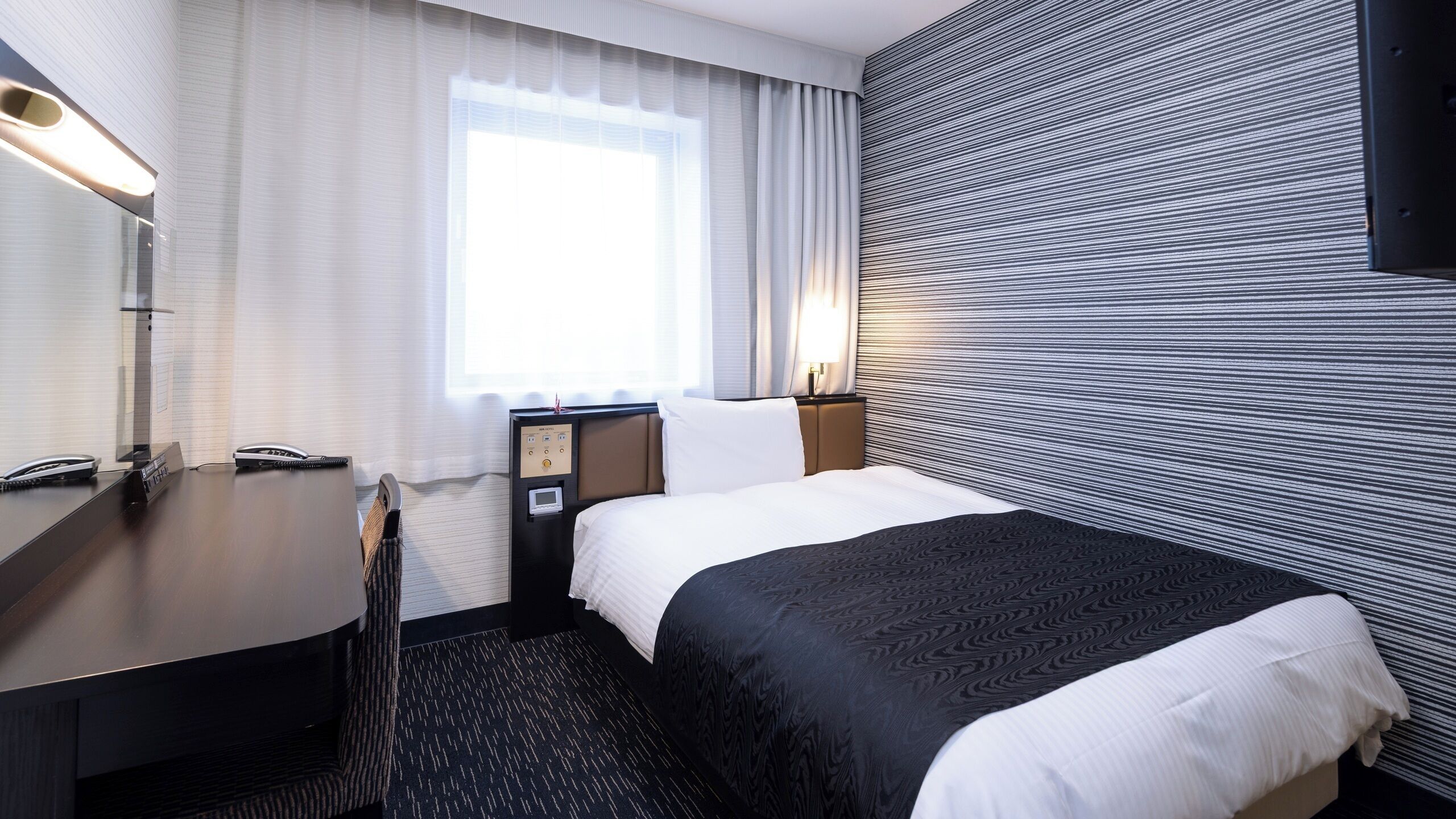 Une chambre d’hôtel équipée d’un lit, d’un bureau, d’un téléphone, d’une table de chevet, d’une lampe et d’une fenêtre avec des rideaux.