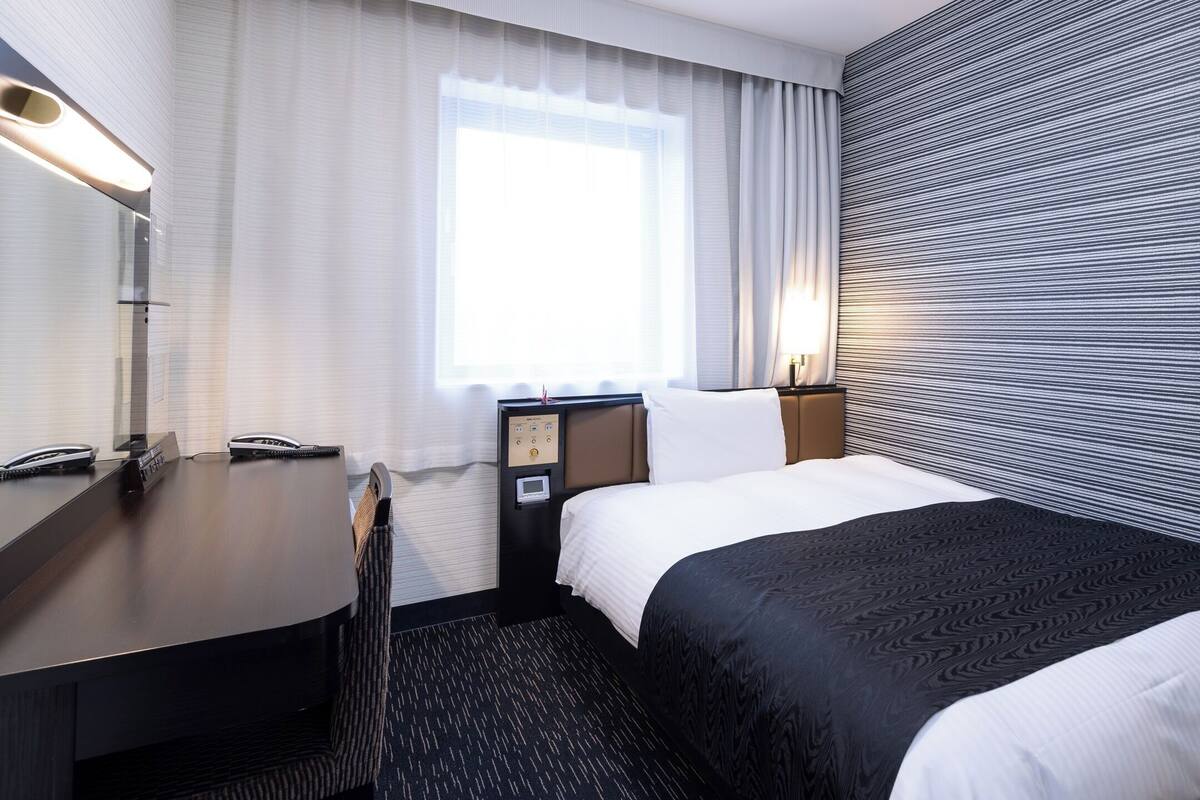 Ein Hotelzimmer mit Bett, Schreibtisch, Telefon, Nachttisch, Lampe und einem Fenster mit Vorhängen.