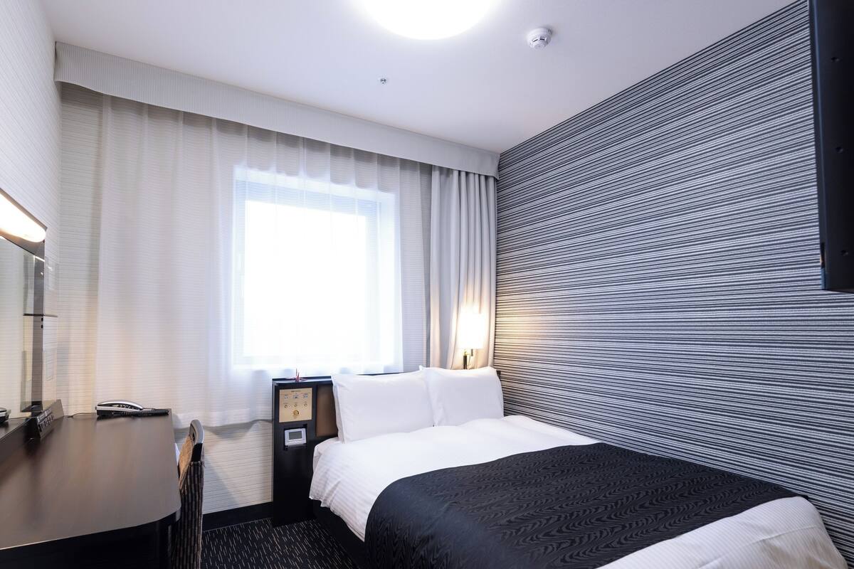 Ein Hotelzimmer mit einem Bett, einem Schreibtisch, einem Stuhl, einem Nachttisch mit Lampe und einem Vorhangfenster.
