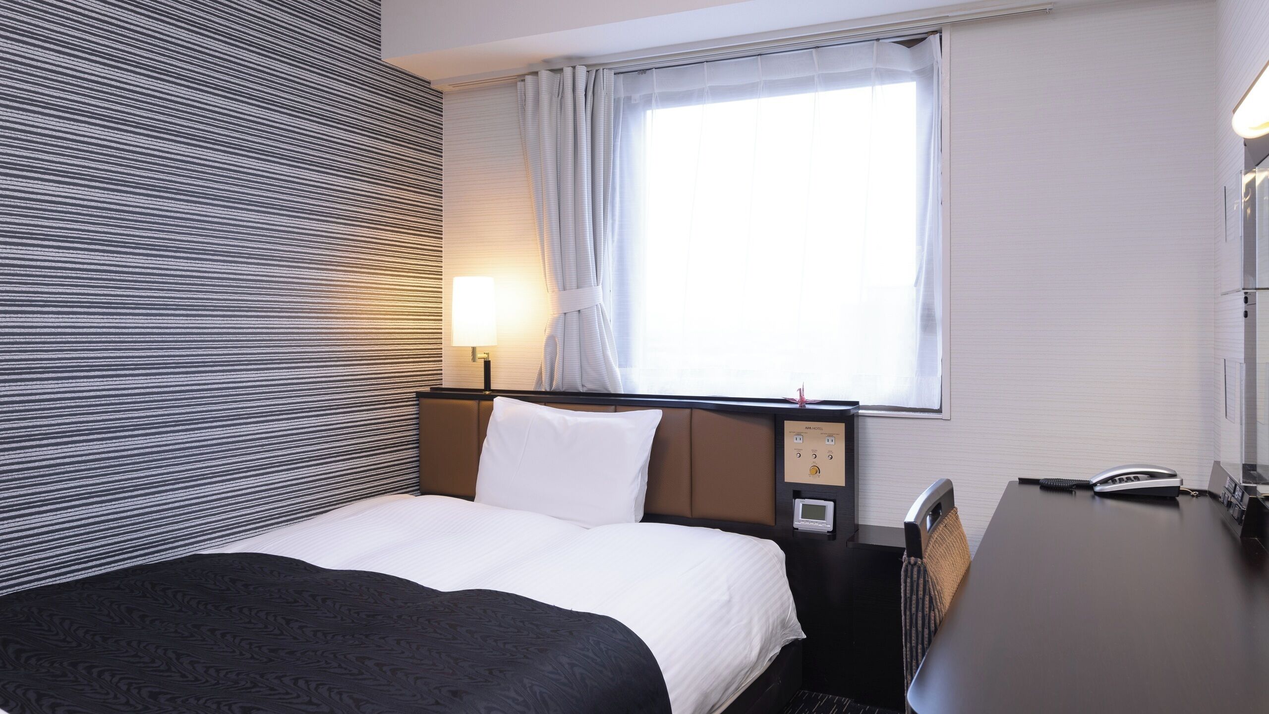 Une chambre d’hôtel comprenant un lit, un bureau, une lampe, une fenêtre avec des rideaux et un mur à motifs rayés.