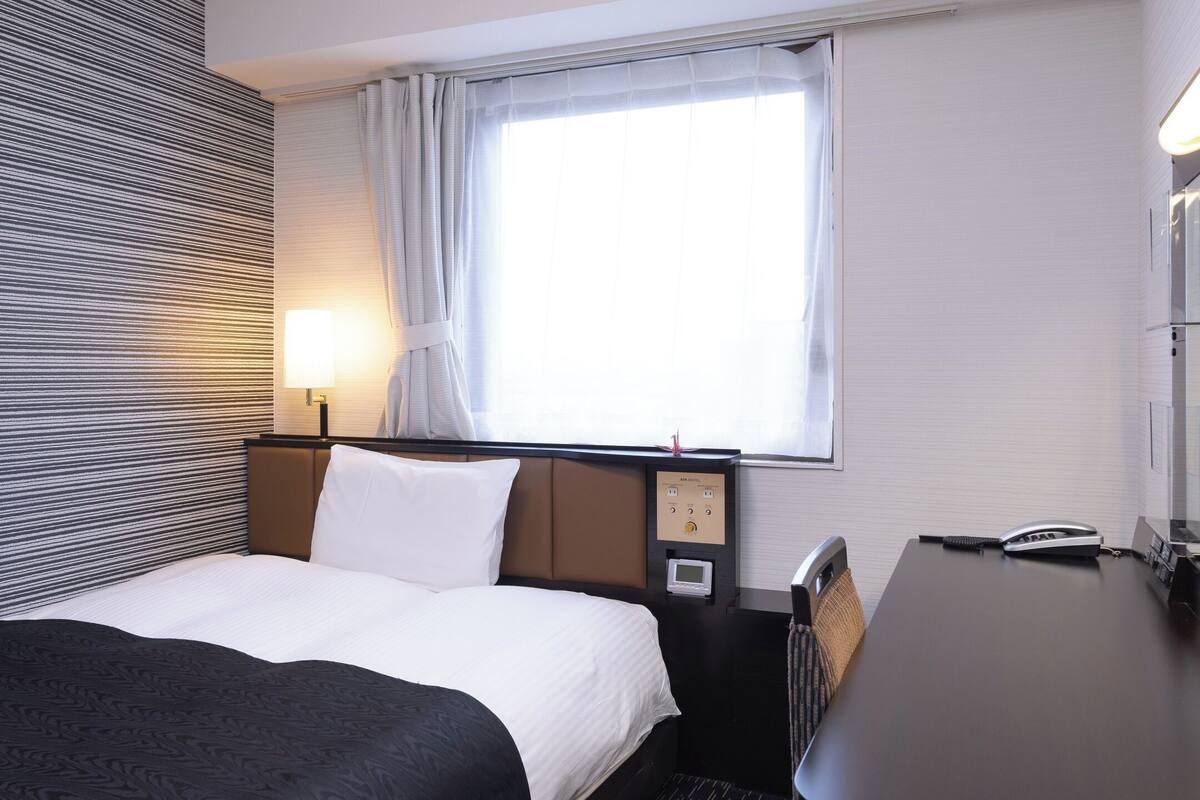 Ein Hotelzimmer mit Bett, Schreibtisch, Lampe, einem Fenster mit Vorhängen und einer Wand mit Streifenmuster.