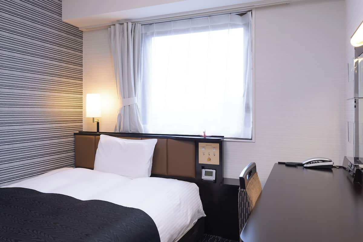 Une chambre d’hôtel comprenant un lit, un bureau, une lampe, une fenêtre avec des rideaux et un mur à motifs rayés.