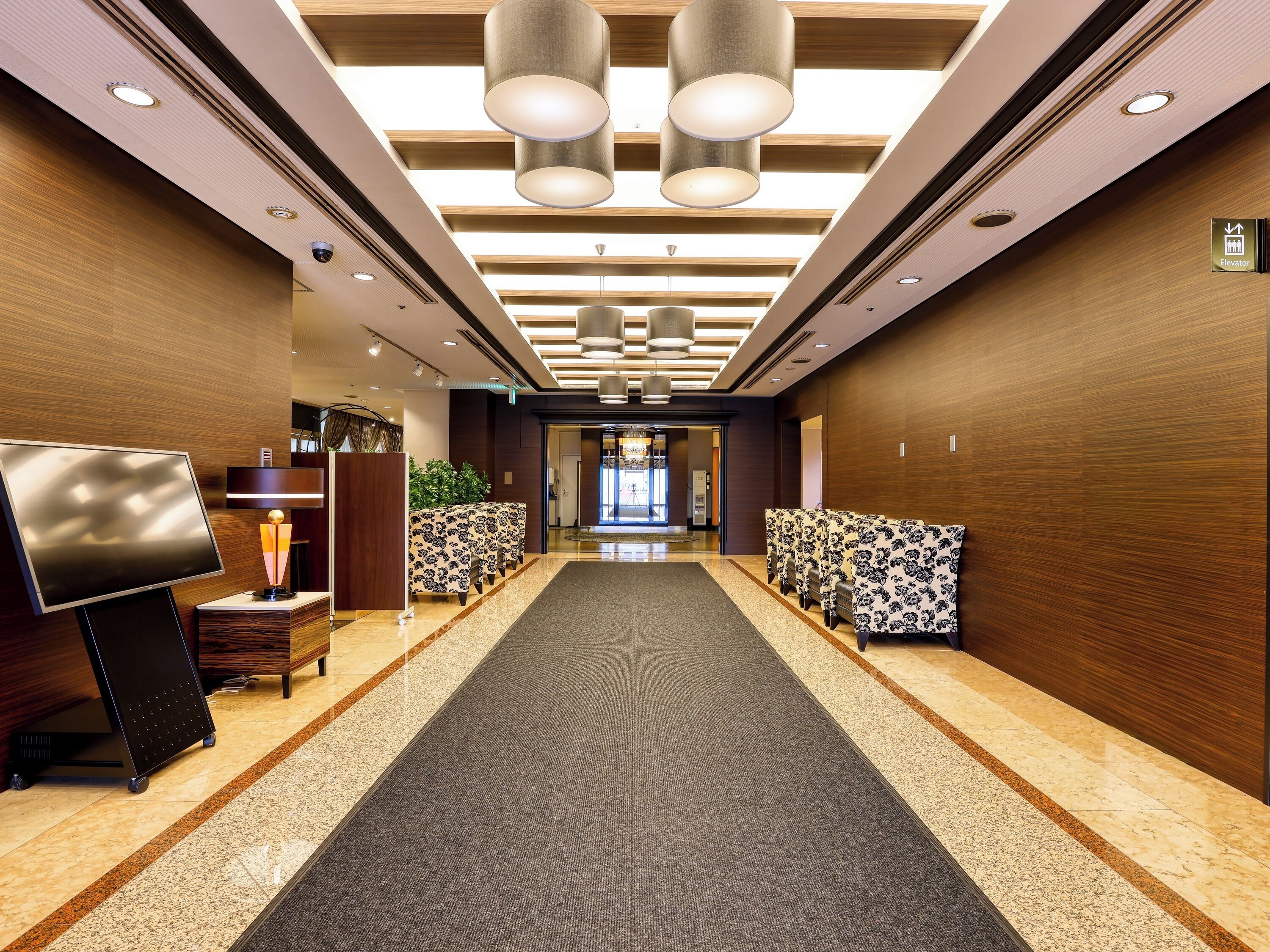 A hotel lobby with a long carpeted corridor, wooden panel walls, and a reception desk with a TV.