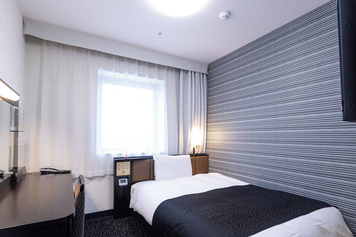 Ein Hotelzimmer mit Bett, Schreibtisch, Stuhl, Nachttisch, Lampe und einem Fenster mit Vorhängen.