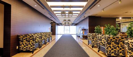 Un hall d’hôtel doté d’un couloir central recouvert de moquette, d’espaces aménagés de chaque côté et d’une grande fenêtre au fond.