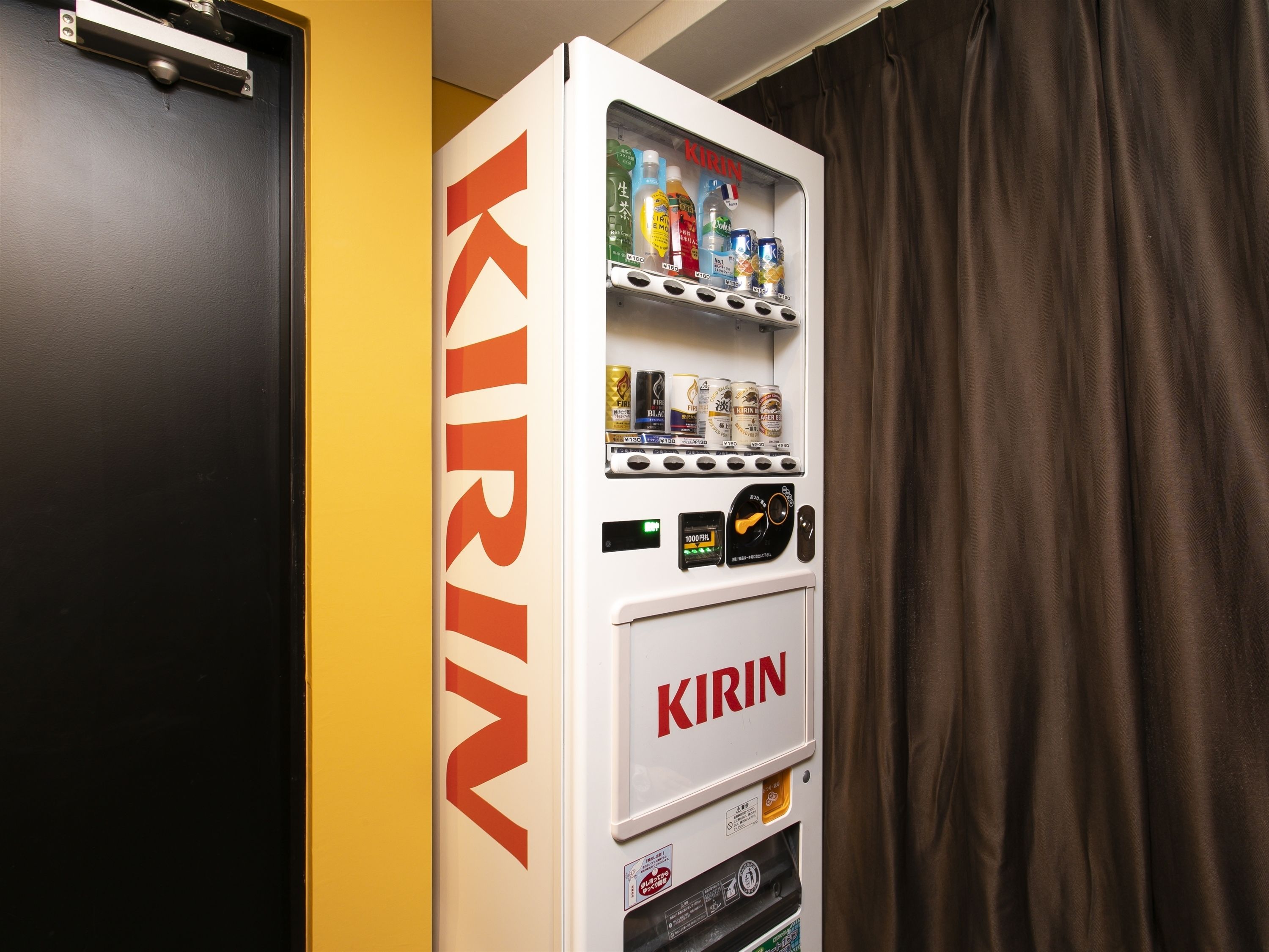 Máquina expendedora con el rótulo "KIRIN" en una habitación con una puerta negra y pared amarilla.
