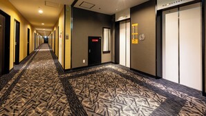 Ein Hotelgang mit einem gemusterten Teppich, Türen an beiden Seiten und einem Aufzug mit digitaler Anzeige.