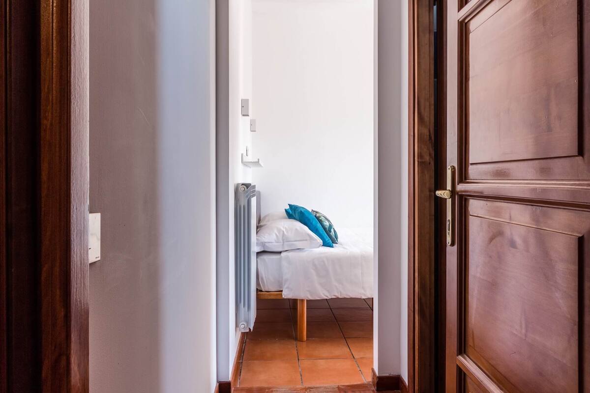A narrow hallway leading to a bedroom with a bed, a radiator, and a door.