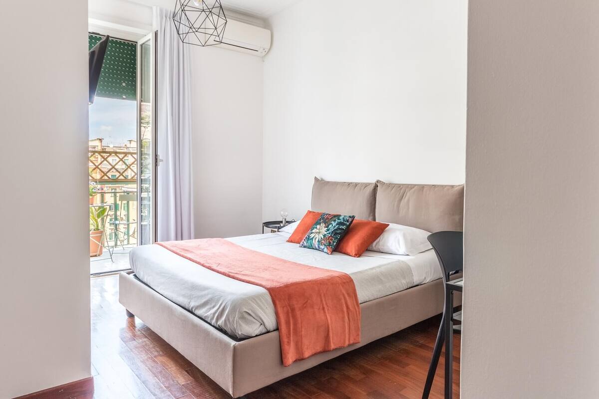 A bedroom with a bed, pillows, a nightstand, a chair, and a view of the outside.