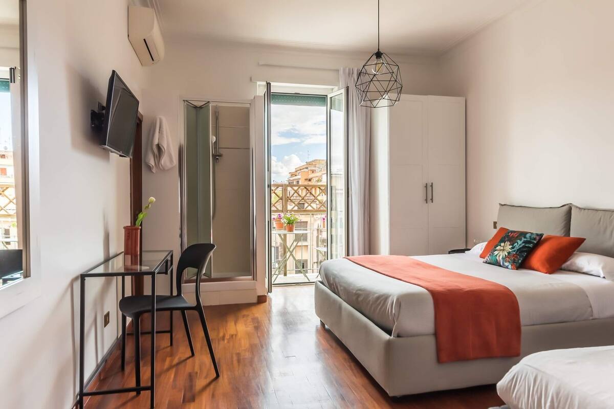 A modern bedroom with a bed, a desk with a chair, a TV, and a balcony with a view of buildings.