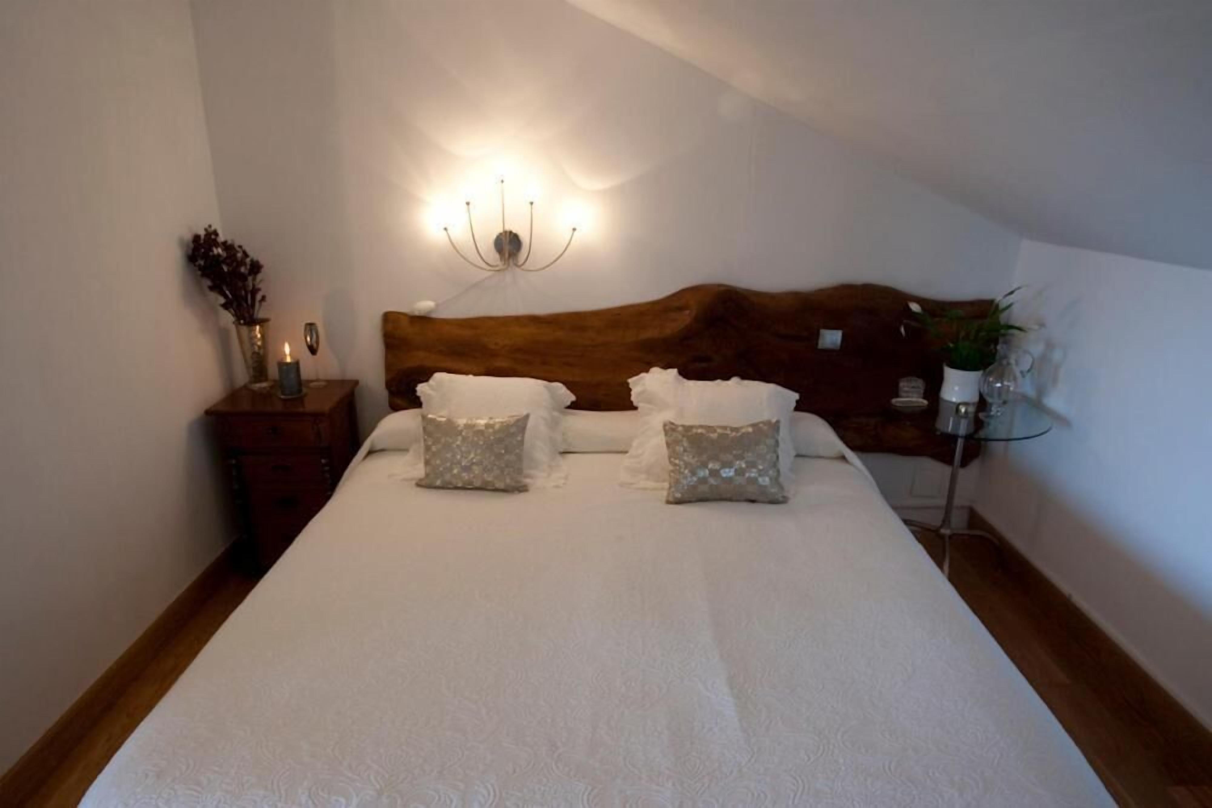 Una cama bien hecha con sábanas blancas y dos almohadones decorativos, una mesita de noche con un florero y una lámpara, y una lámpara de pared.