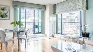 Una sala de estar moderna con una mesa de vidrio, sillas blancas, un sofá y una mesa de centro. Amplias ventanas ofrecen vistas a un paisaje urbano.
