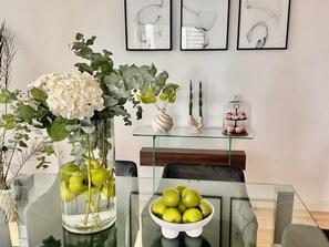 Une table à manger ornée d’un vase en verre contenant des fleurs blanches et des pommes vertes, d’un bol rempli de pommes vertes et d’une vitrine où sont exposés des objets décoratifs.