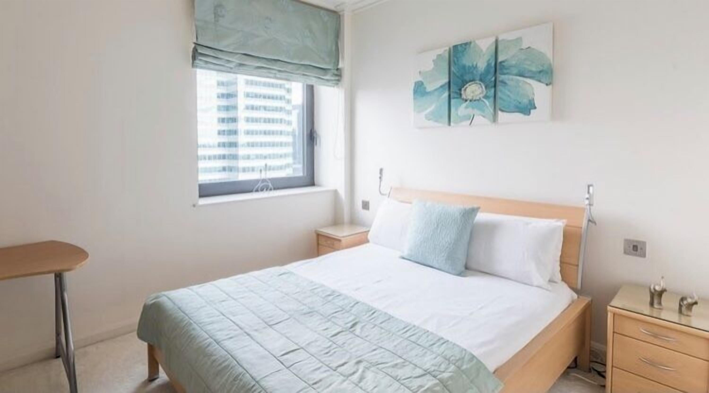 Un dormitorio con cama, mesita de noche, ventana con persianas y una obra de arte colgada en la pared.