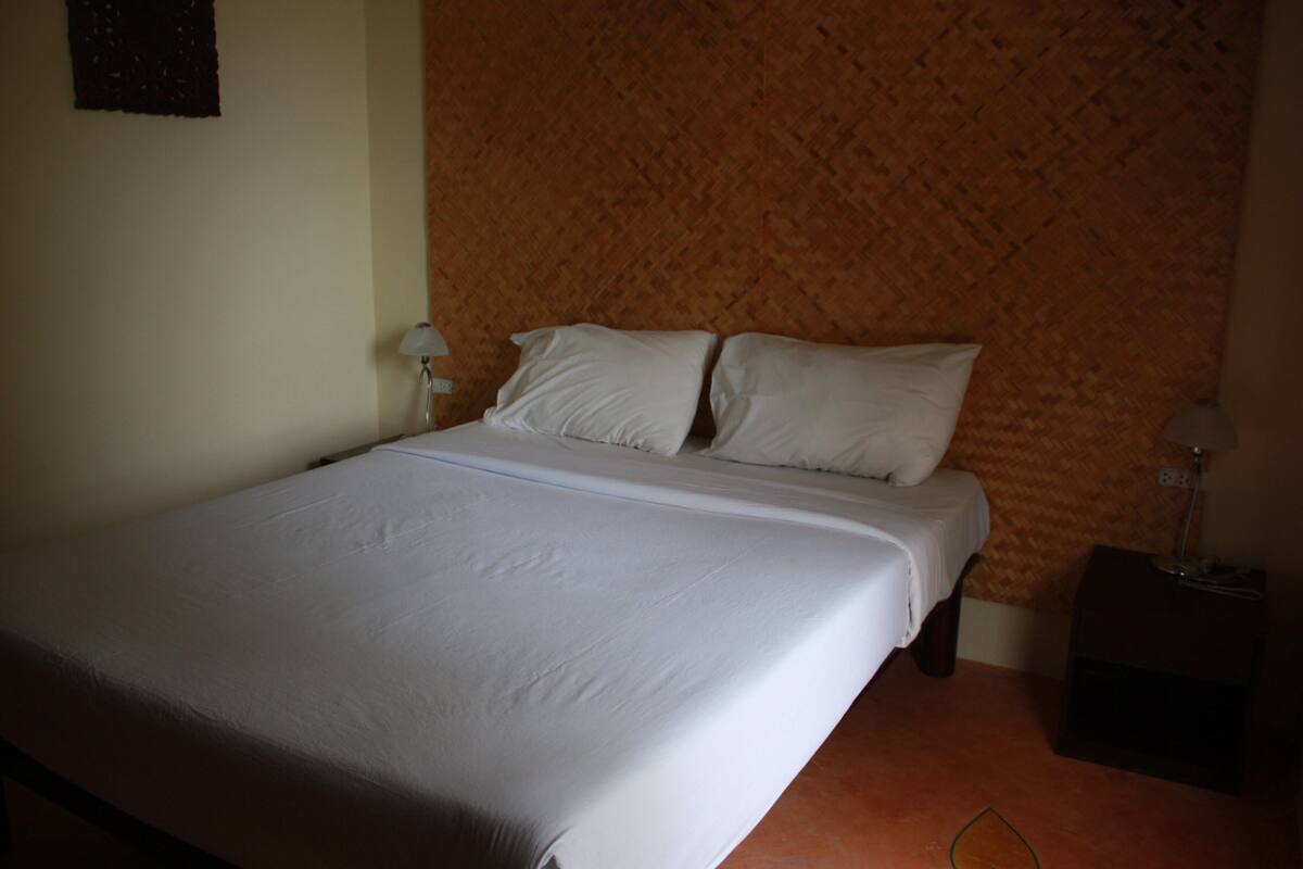 Un lit bien fait, avec du linge de lit blanc, dans une pièce dont un mur est peint dans une nuance d’orange texturée, et sur laquelle se trouve une petite table de chevet avec une lampe.