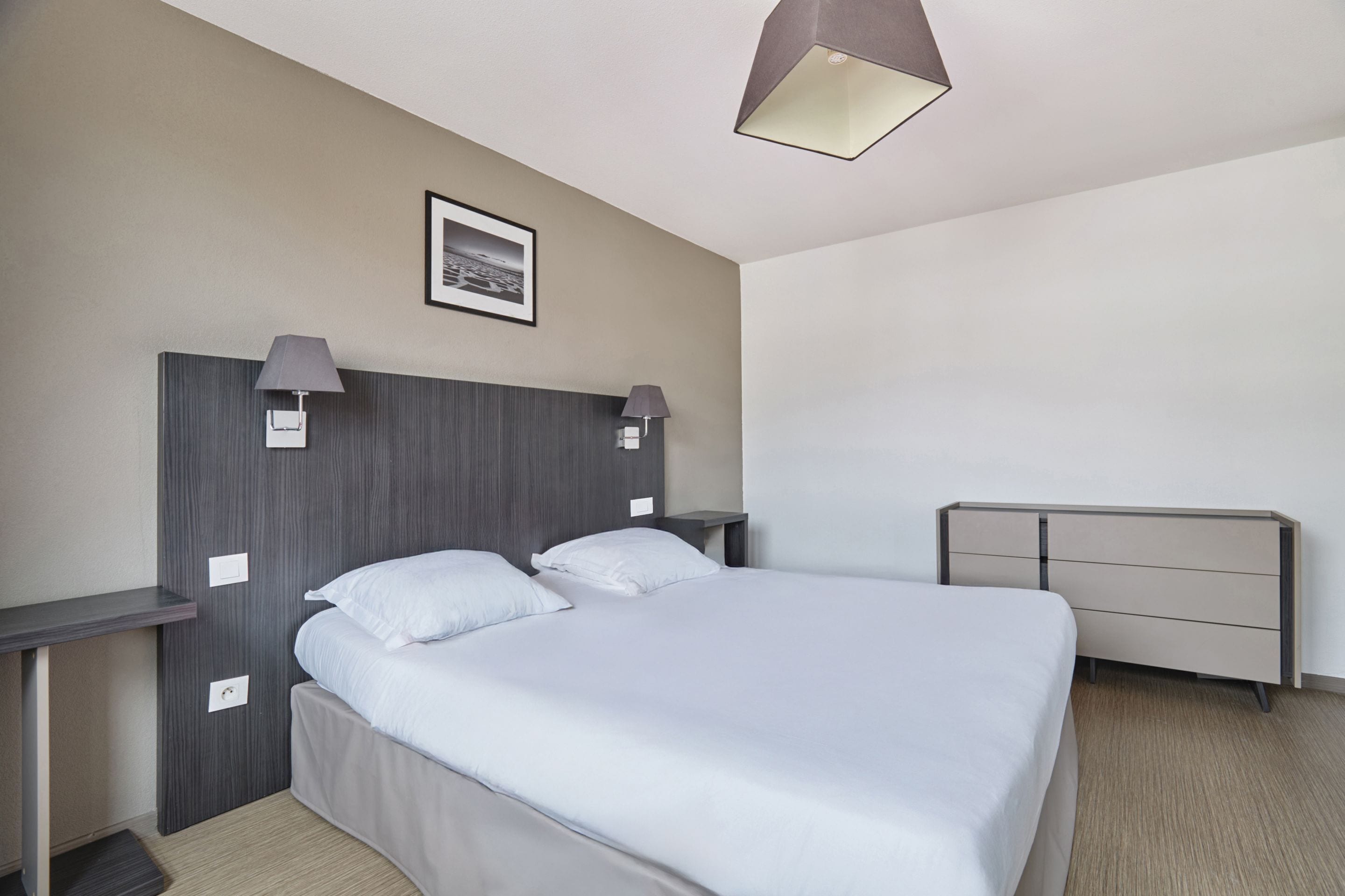 A modern hotel room with a large bed, two bedside tables, a nightstand, a headboard, a wall-mounted lamp, and a framed picture.