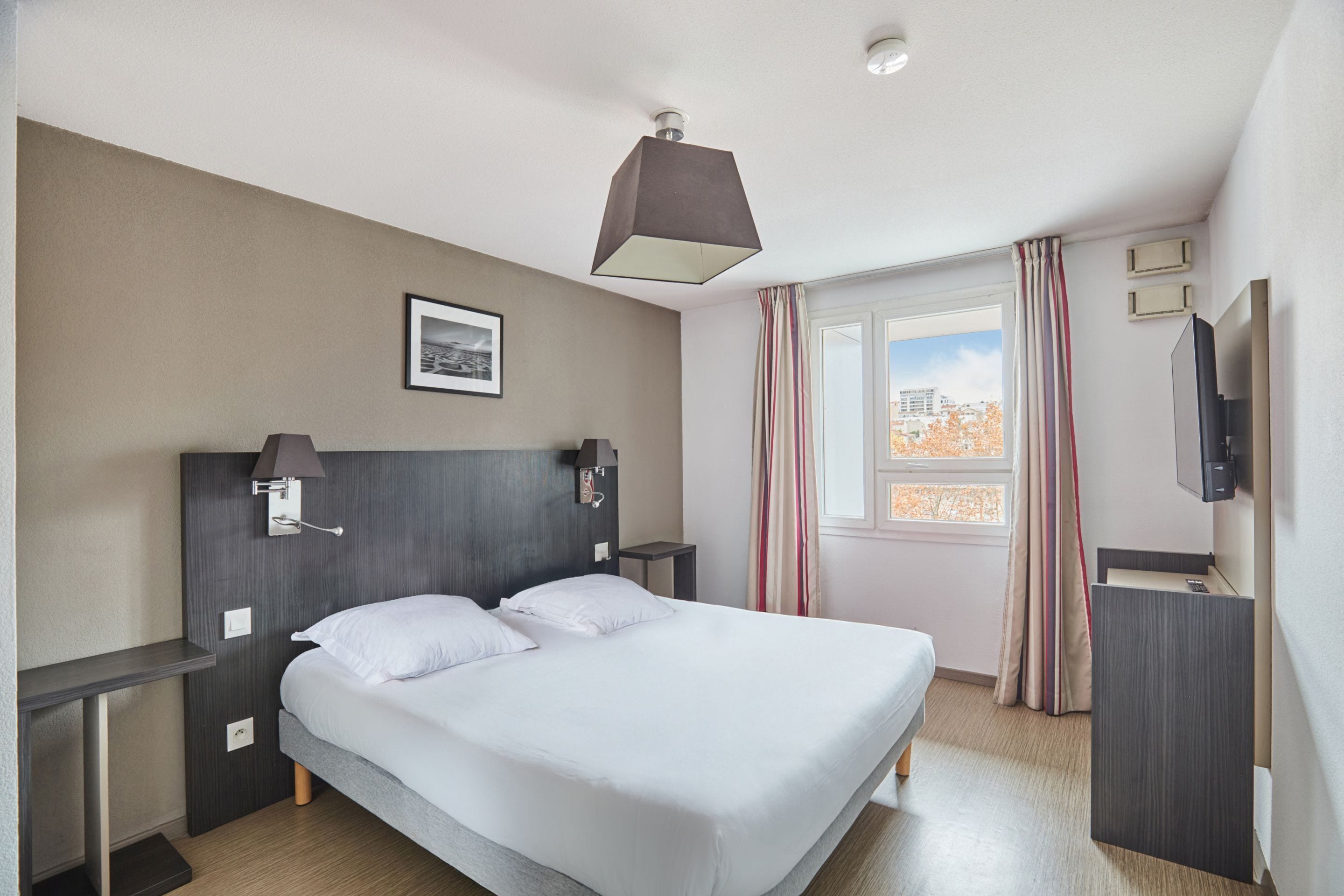 A hotel room with a large bed, bedside tables, a flat-screen TV, a window with curtains, and a ceiling light fixture.