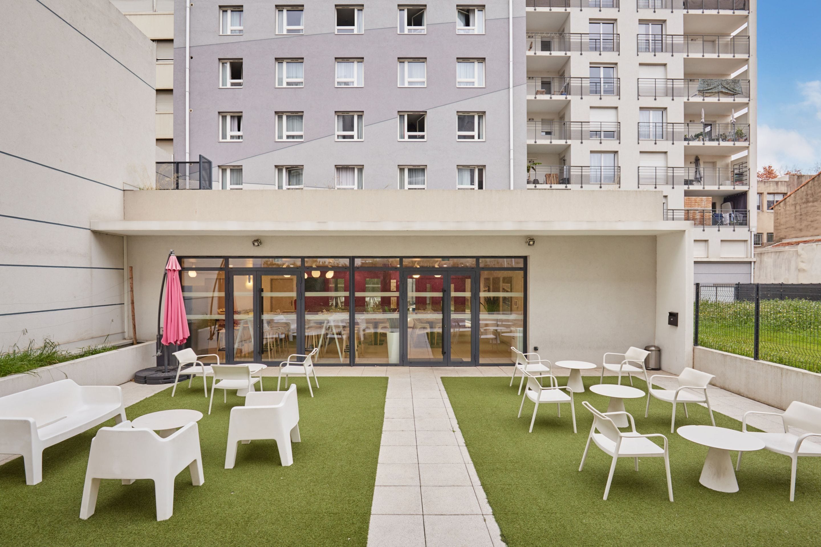 Ein Innenhof mit Außenbestuhlung, ein Glastür-Eingang und ein Gebäude mit mehreren Balkonen im Hintergrund.