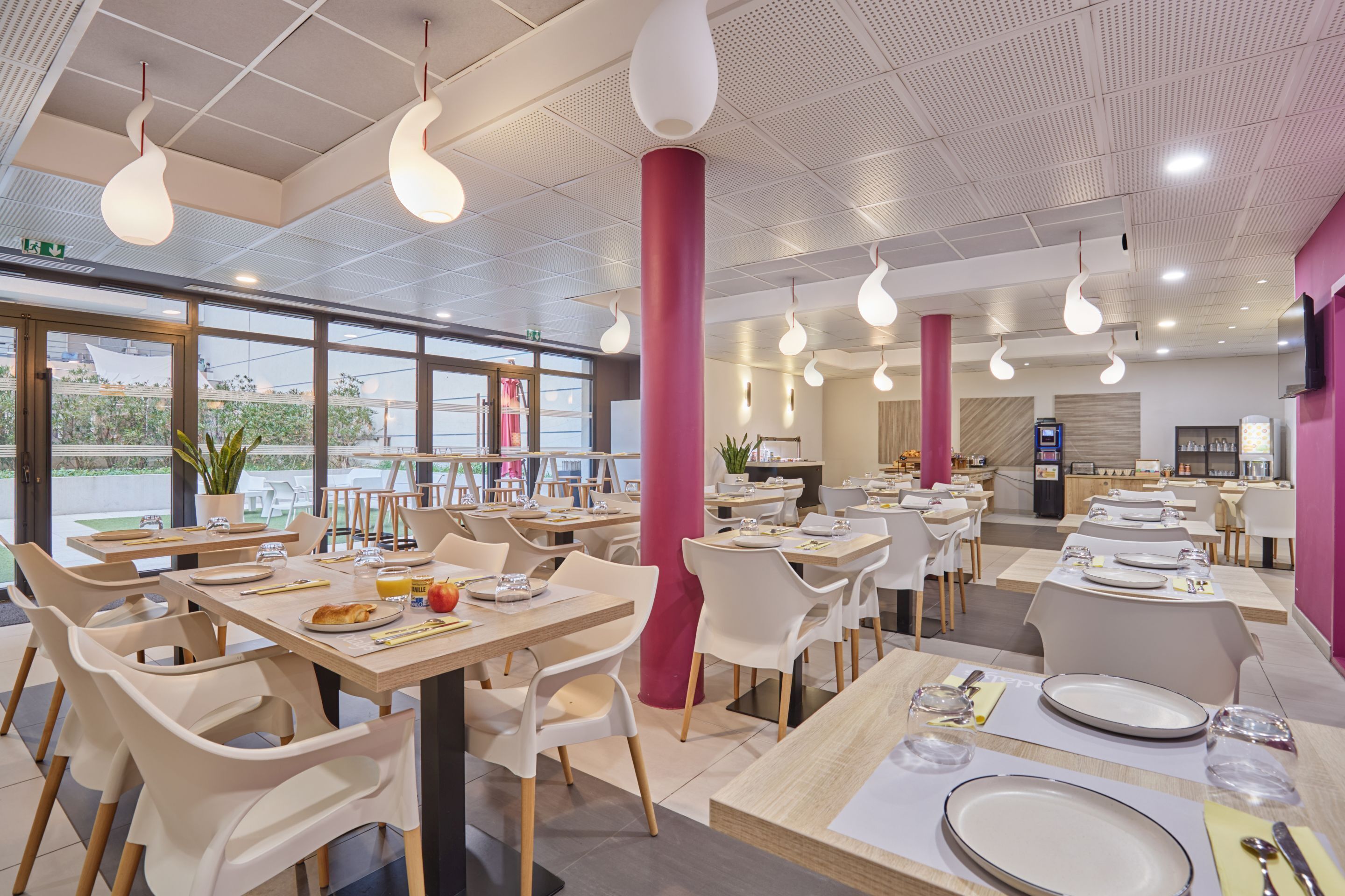 A modern restaurant with white chairs and tables, a pink column, and hanging light fixtures.