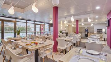 Un restaurant moderne avec des chaises et des tables blanches, une colonne rose et des luminaires suspendus.