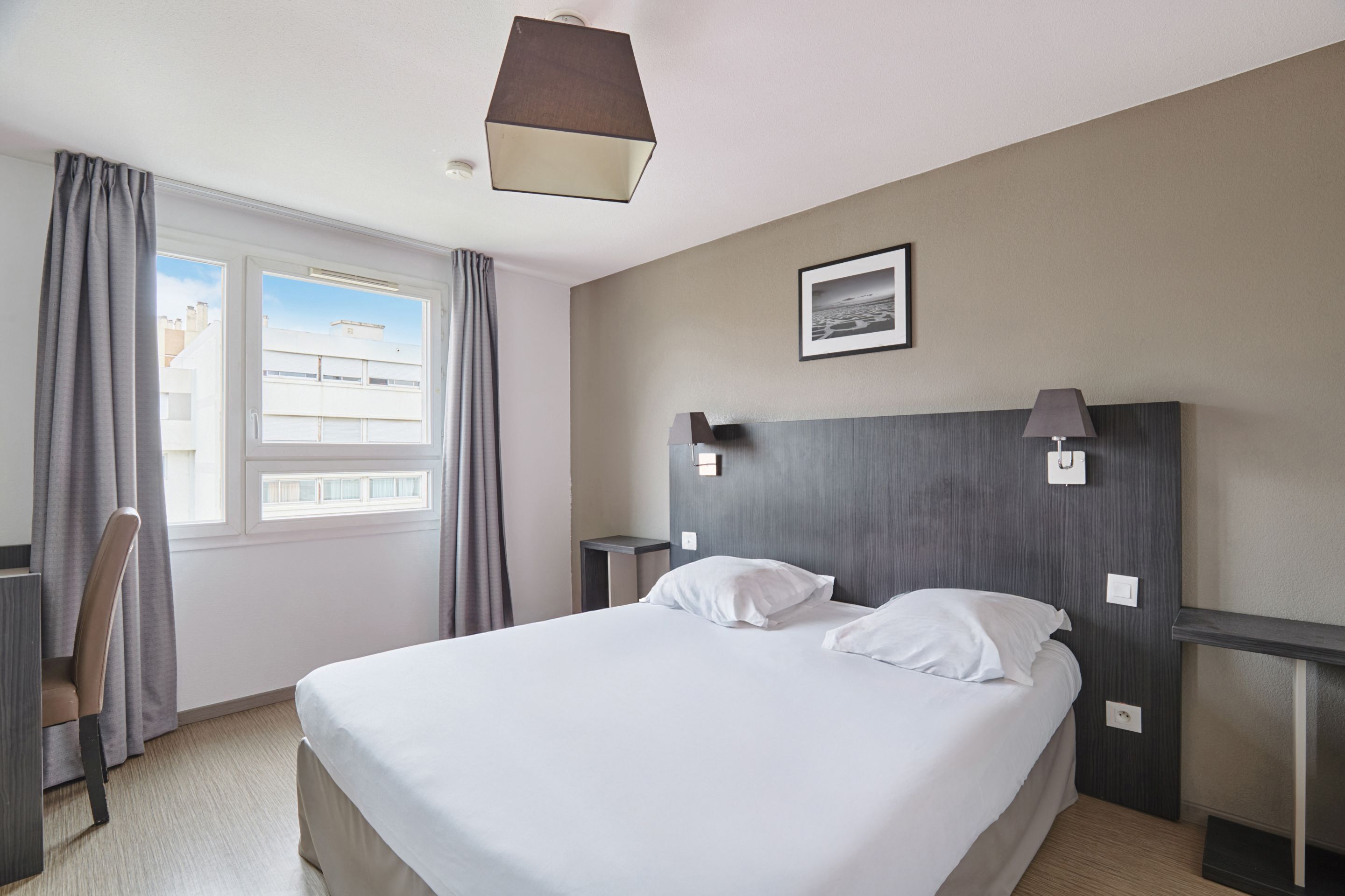 Une chambre d’hôtel avec un grand lit, un bureau, une chaise, une fenêtre avec des rideaux, un tableau au mur et une lampe sur la table de chevet.