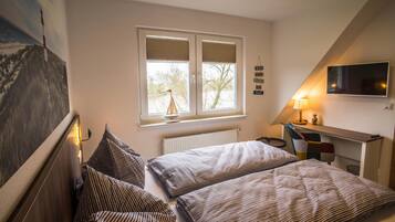 A bedroom with two beds, a window with blinds, a desk with a chair, a TV mounted on the wall, and a radiator.