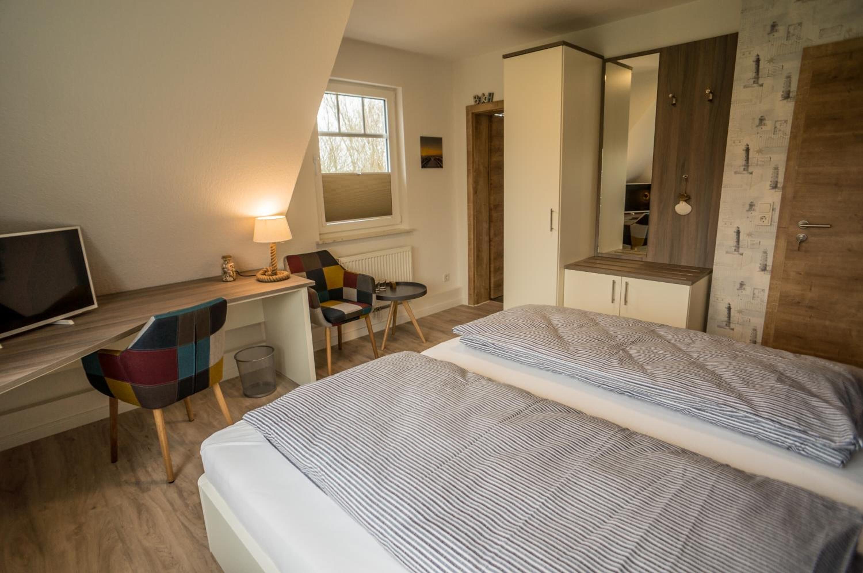 A bedroom with a bed, a desk with a chair, a TV, a window, and a wardrobe.