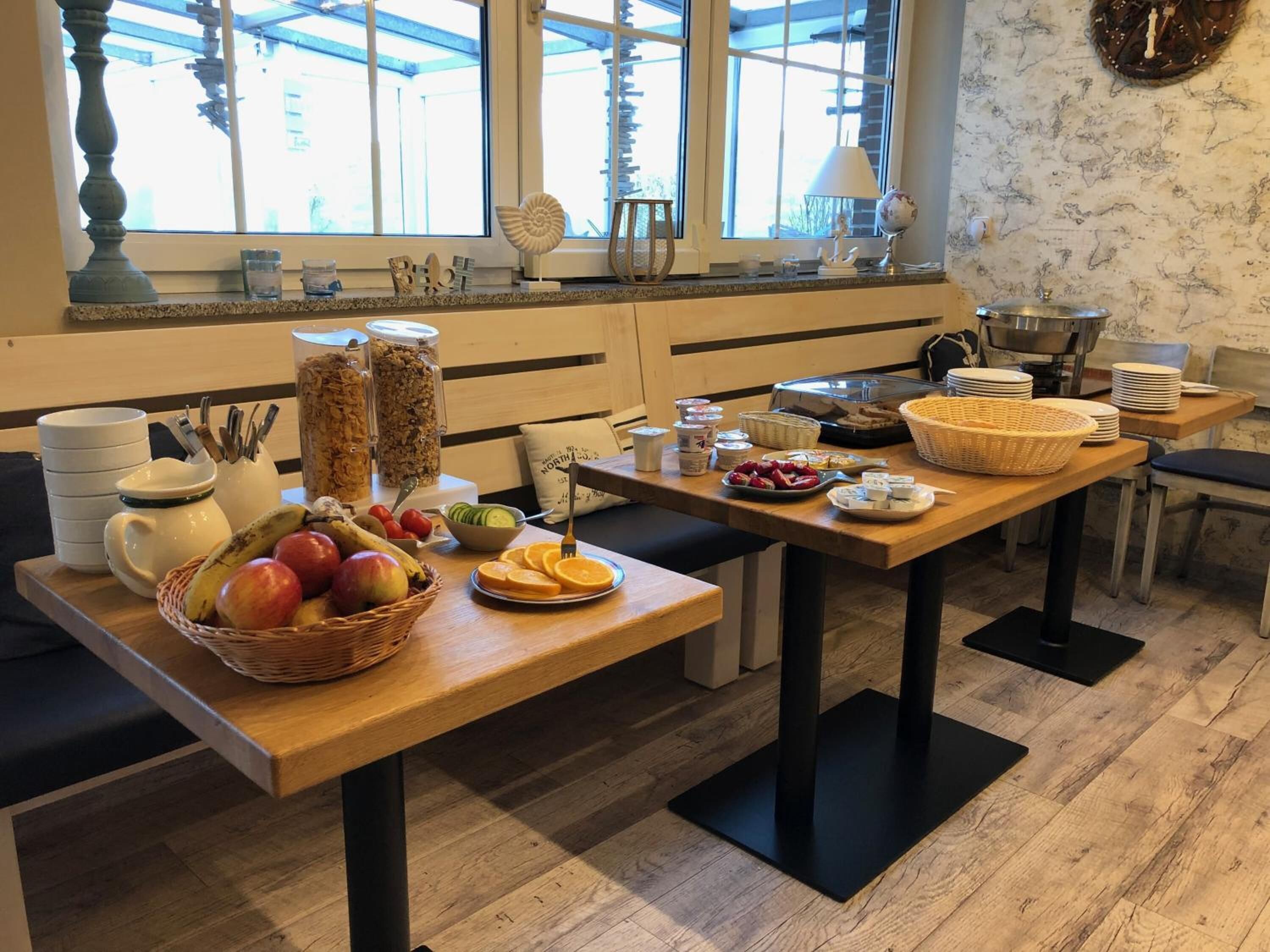 A breakfast buffet setup with various food items and utensils on a wooden table, with a view of the outside through large windows.