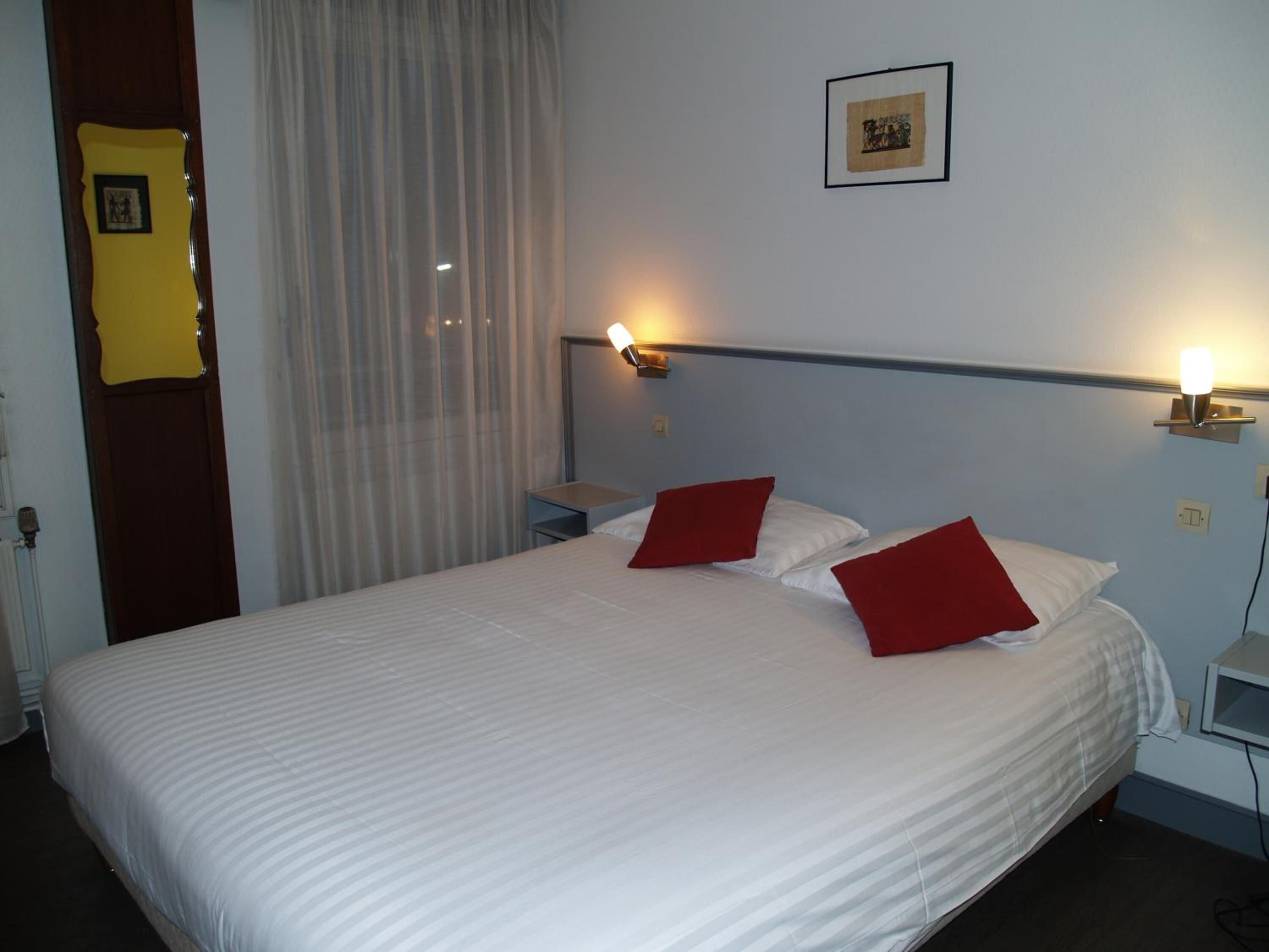 Une chambre d’hôtel avec un grand lit, deux oreillers rouges, une table de chevet, une lampe fixée au mur et un tableau encadré accroché au mur.
