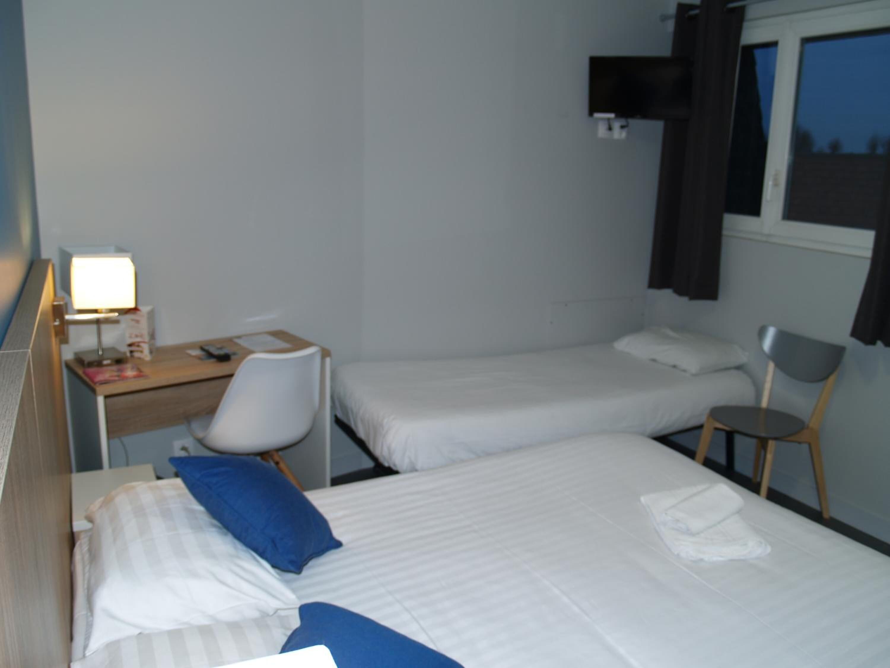 A hotel room with two beds, a desk with a chair, a lamp, and a window with curtains.