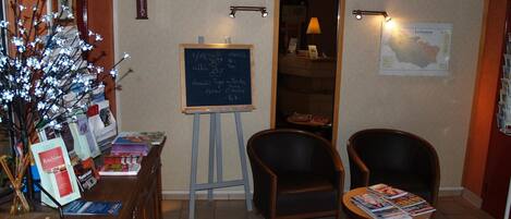 A reception area with a desk displaying books and magazines, a blackboard with handwritten notes, two chairs, and a wall map.