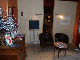 Zona de recepción con un mostrador que exhibe libros y revistas, una pizarra con notas escritas a mano, dos sillas y un mapa mural.