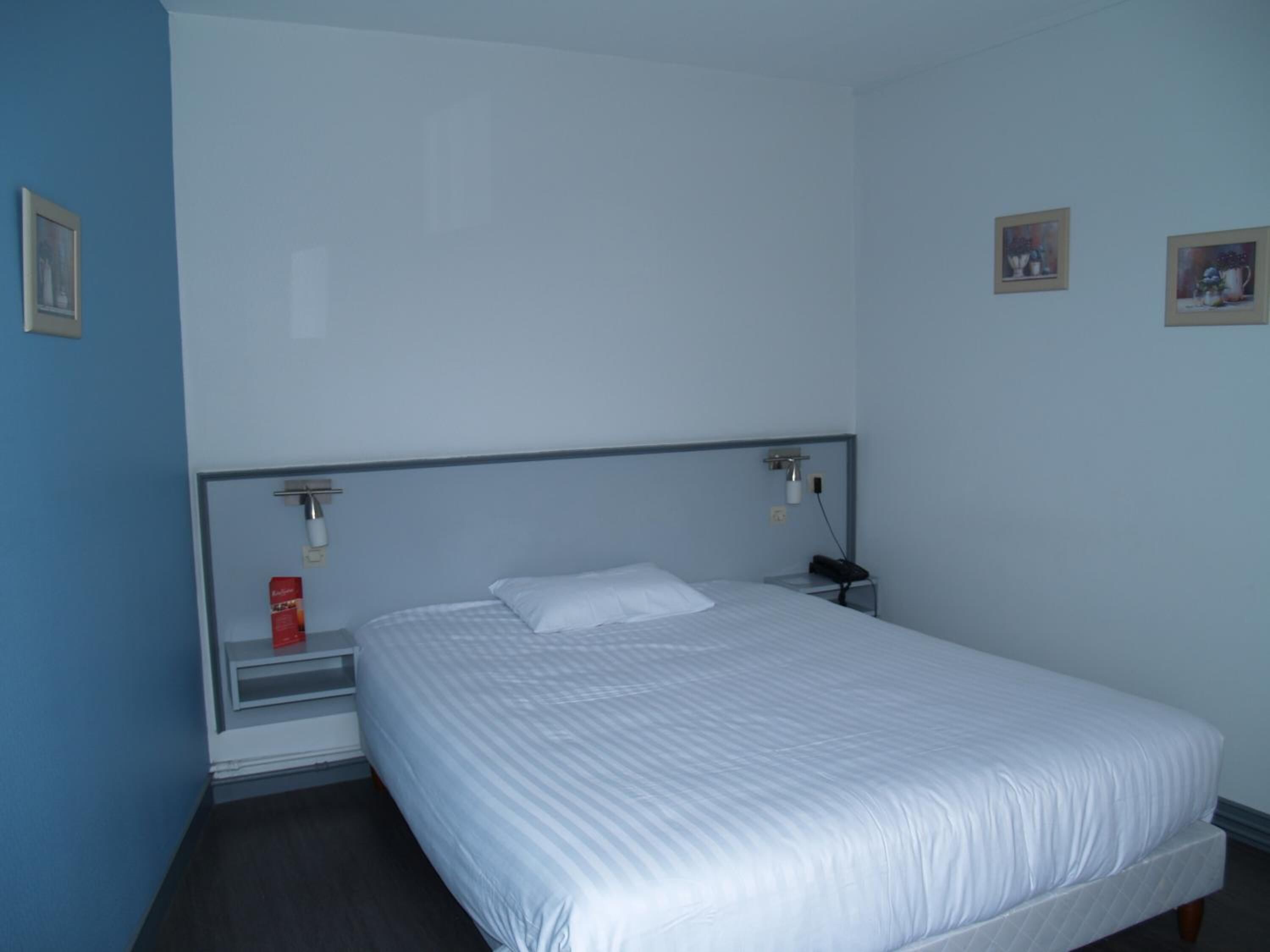 Ein Zimmer mit einem Einzelbett, einem Holzkopfteil, einem Nachttisch mit Lampe und zwei gerahmten Bildern an der Wand.