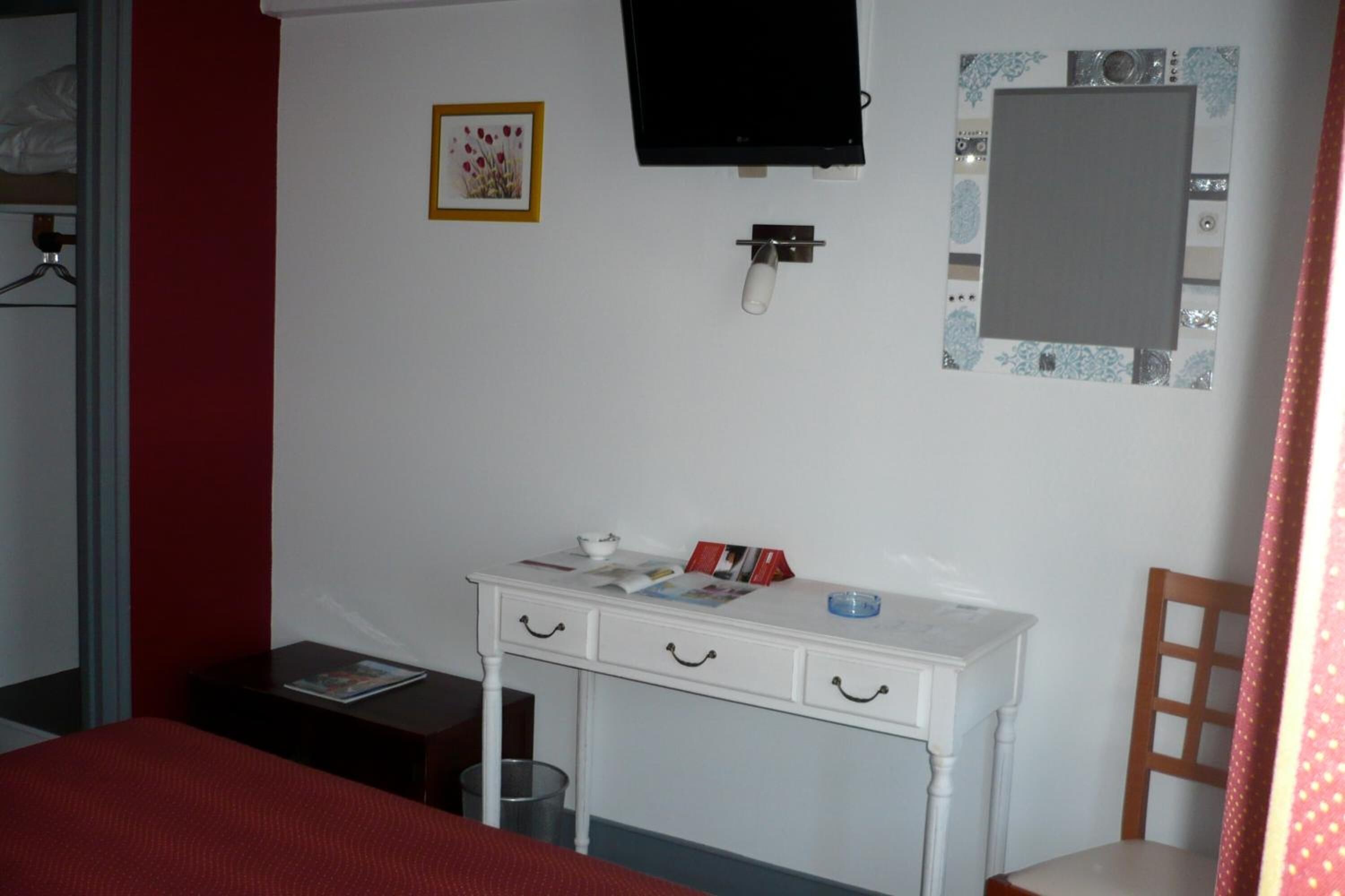 Une chambre à coucher avec un lit, un bureau blanc avec des tiroirs, une chaise, un téléviseur fixé au mur et une petite poubelle.