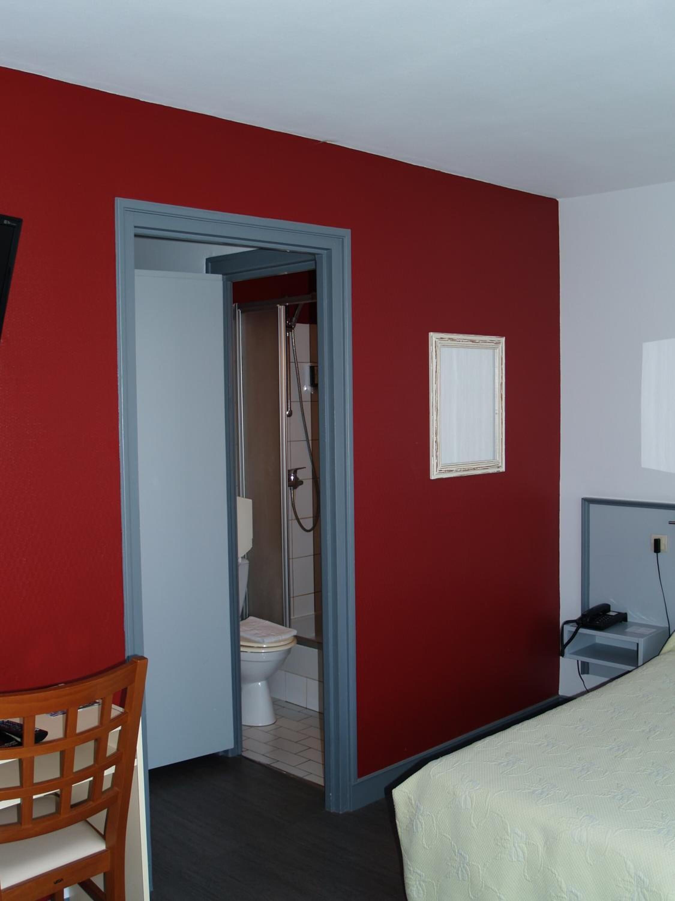 Een hotelkamer met een rode wand, een witte deur naar een badkamer, een houten stoel, een bed en een klein nachtkastje met een lamp.