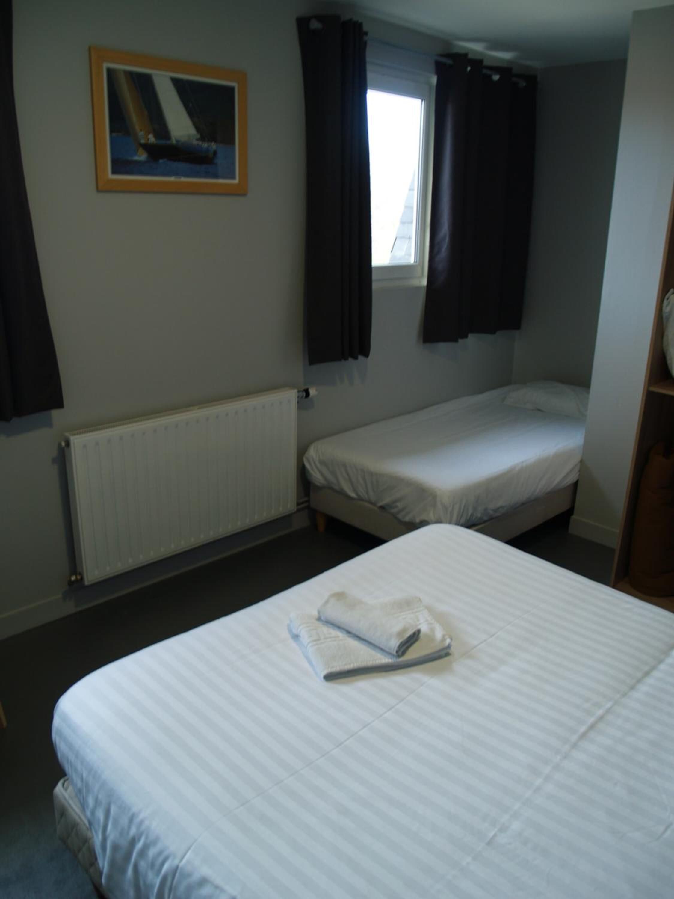 A small, single-room hotel room with a bed, a window with curtains, a radiator, and a framed picture on the wall.