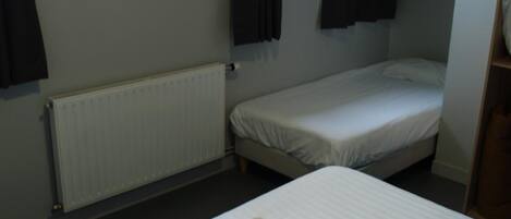 A small, single-room hotel room with a bed, a window with curtains, a radiator, and a framed picture on the wall.