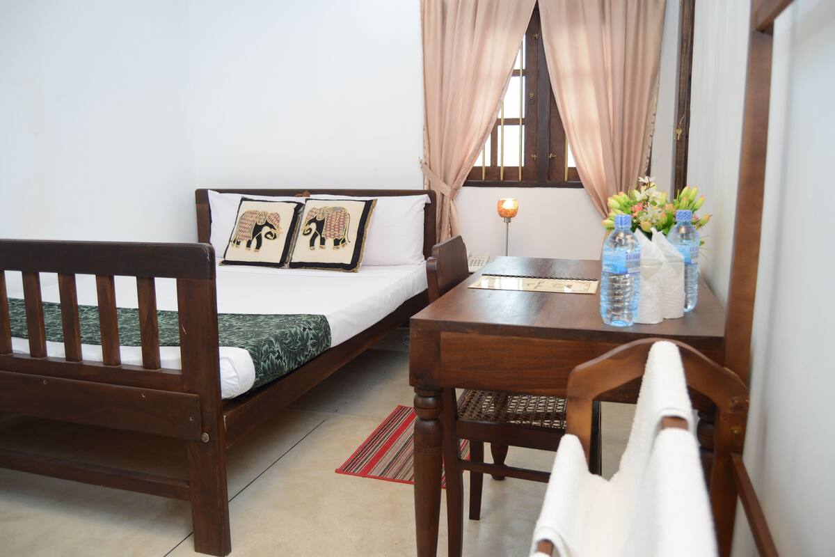 A small, cozy room with a bed, a wooden chair, a table, and a mirror.
