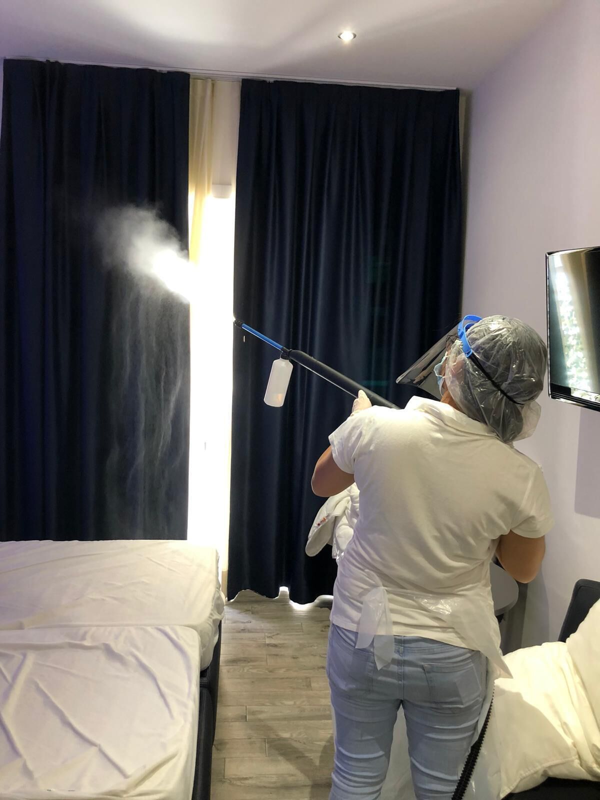 A person cleaning a window with a spray bottle, a bed, and a TV mounted on the wall.