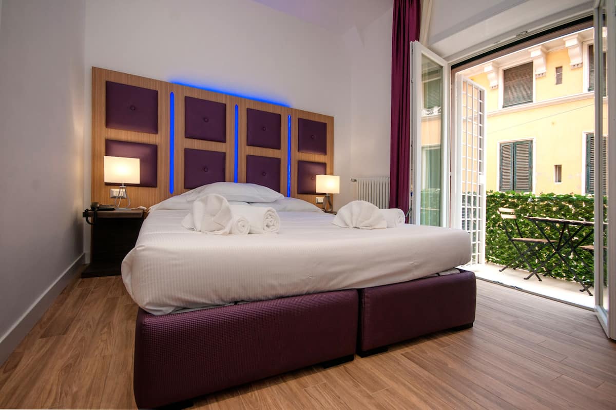 A hotel room with a large bed, two bedside tables with lamps, a wooden headboard with illuminated panels, and a view of a courtyard through a glass door.