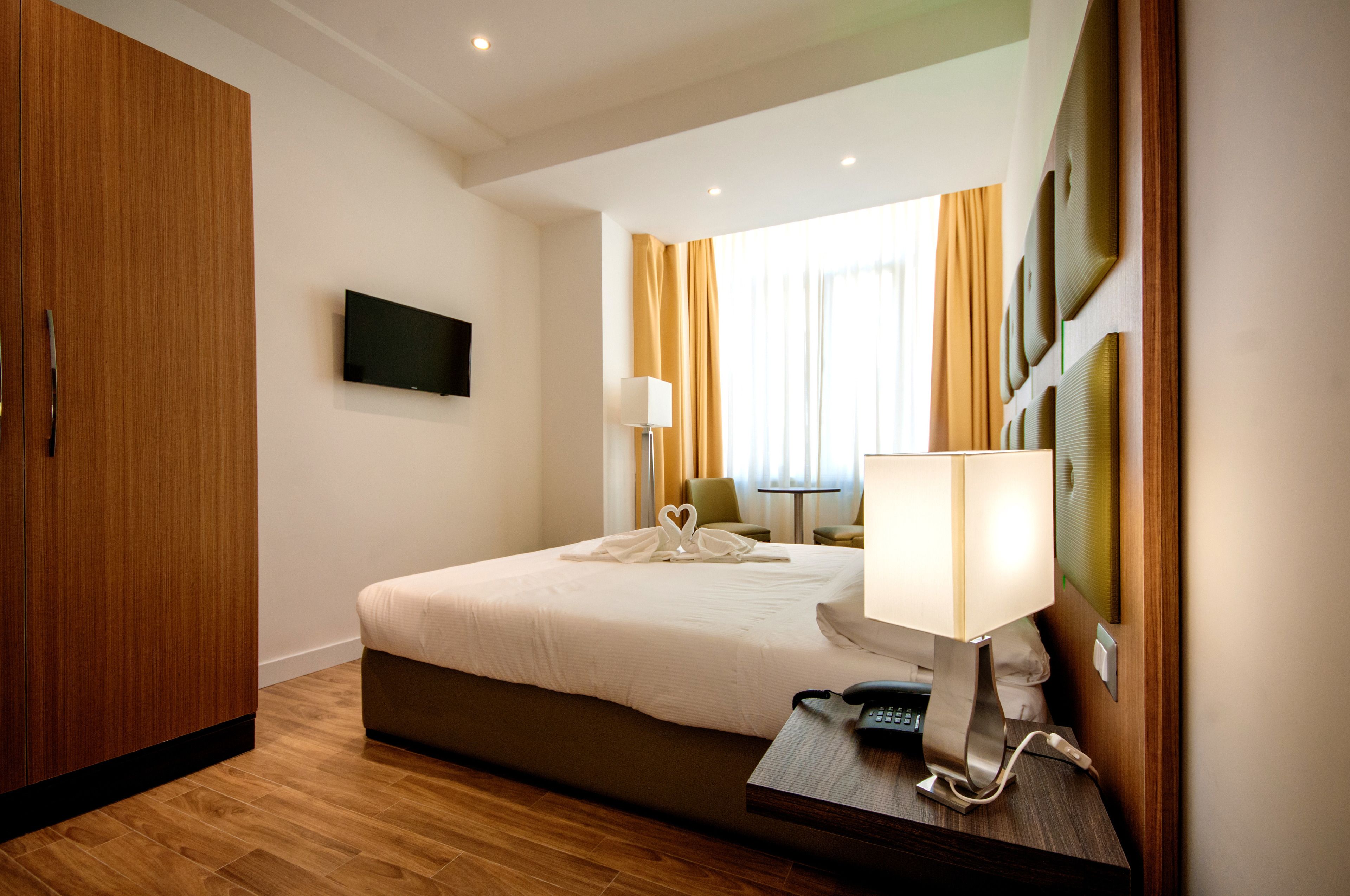 A hotel room with a bed, a desk with a lamp, a telephone, a television on the wall, and a wardrobe.