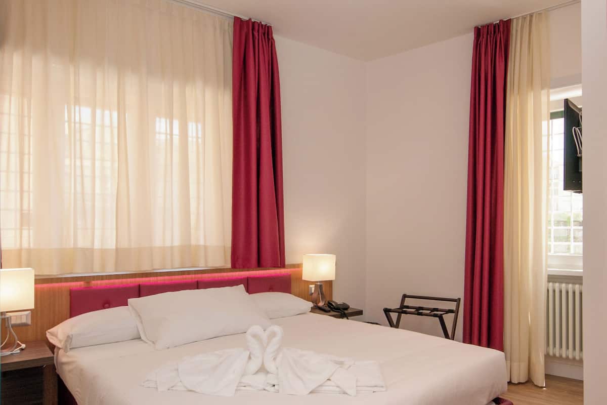 A hotel room with a large bed, a bedside table with a lamp, a chair, and a view of the outside.