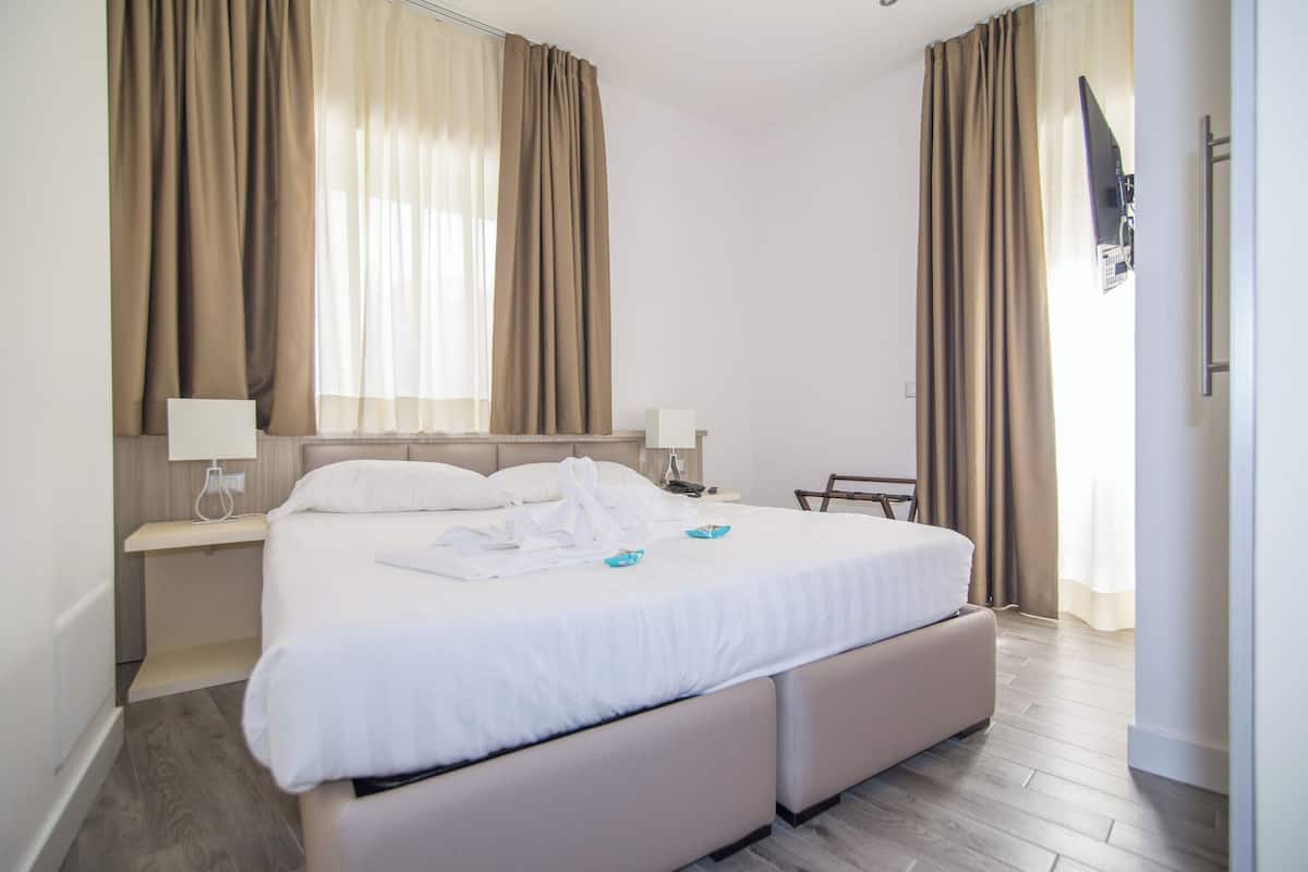 A hotel room with a large bed, bedside tables, a TV mounted on the wall, and a window with curtains.