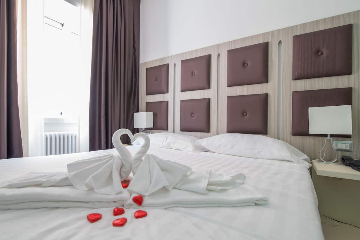 A hotel room with a neatly made bed, a headboard with square panels, a bedside table with a lamp, and a view of the outside through a window with curtains.