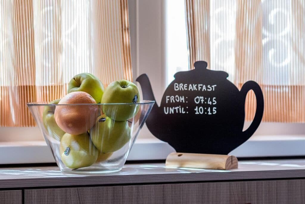 Un tazón de vidrio transparente con manzanas verdes y un melocotón, junto a una tetera negra con asa de madera y un letrero que indica los horarios del desayuno.