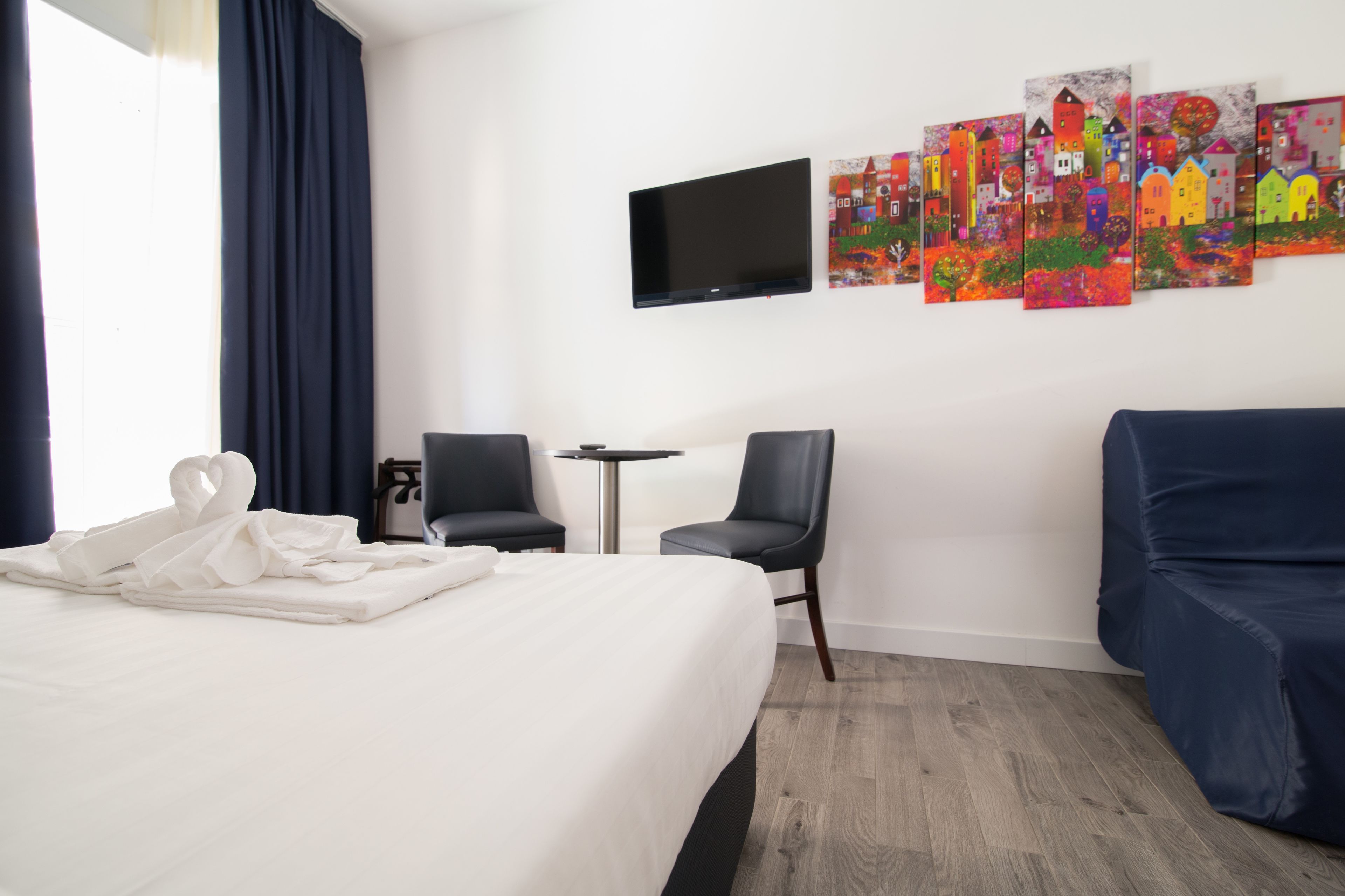 Une chambre d’hôtel avec un lit, deux chaises, une petite table, un téléviseur et des œuvres d’art colorées accrochées au mur.