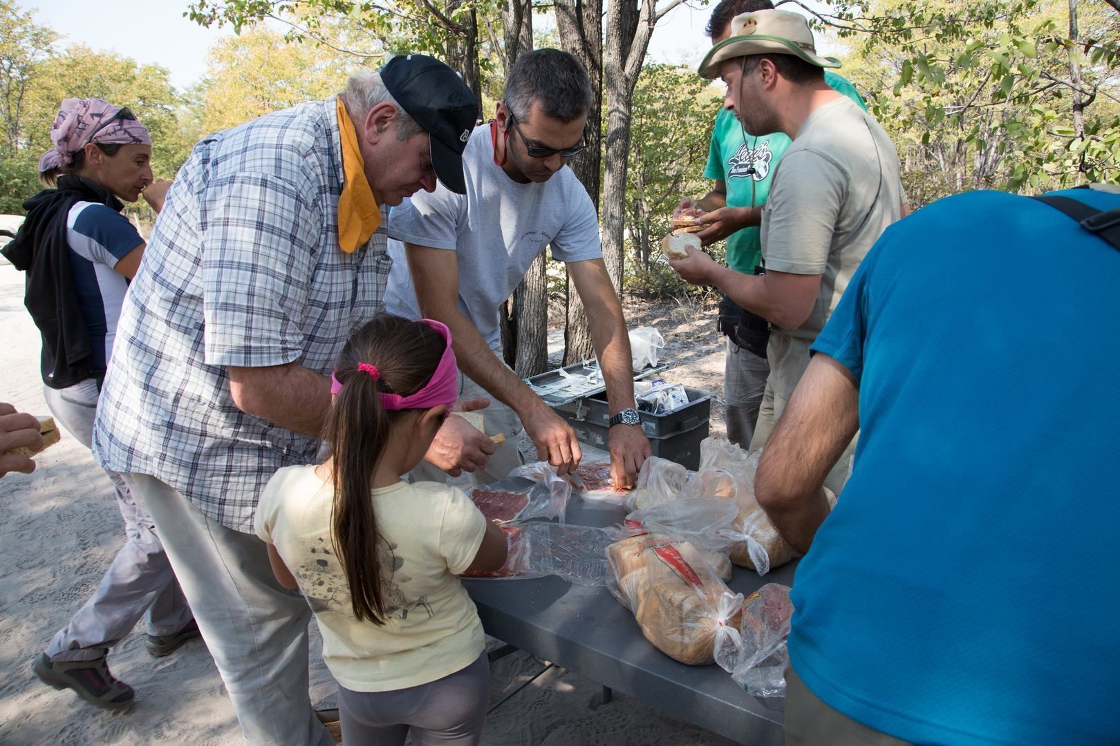 A group of people gathered around a table with food items, possibly at an outdoor event or gathering.