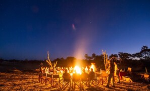 A group of people gathered around a campfire in an open field at night. - Xakanaka Mobile Camp (Xakanaxa)