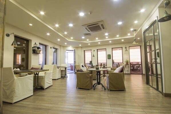 A spacious indoor area with multiple seating arrangements, including sofas and chairs, a central table, and a dining table. The room has a high ceiling with recessed lighting and large windows allowing natural light. - Velvet Hotel (Tbilisi)
