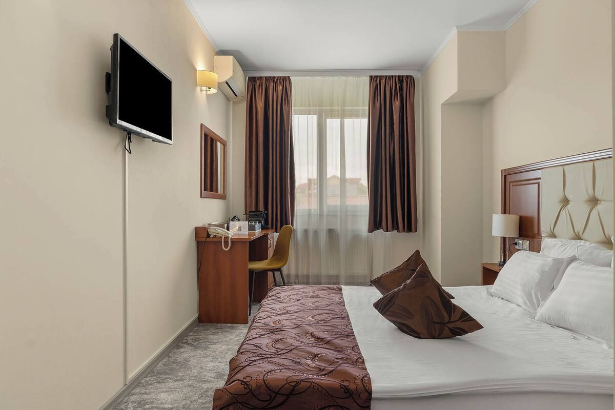 A hotel room with a bed, a desk with a chair, a television, and a window with curtains.